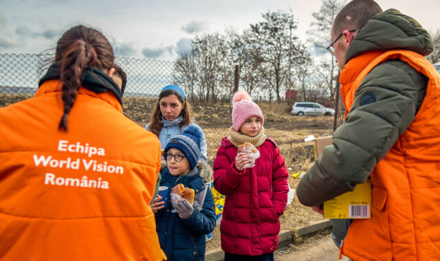 Three children wearing winter coats and hats and holding baked goods and treats face two World Vision staff members in heavy orange vests, a woman on the left and a man on the right.
